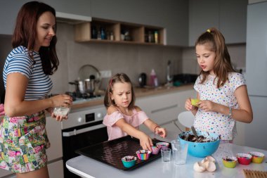 Bir anne ve iki kızı neşeli bir şekilde kek pişiriyorlar. Parlak, modern bir mutfakta. Aile, ortak yemek pişirme deneyiminin eğlenceli ve yaratıcılığını seviyor..