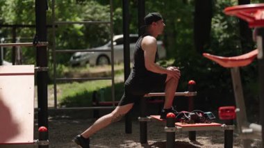 Genç bir atletik adam dışarıda bir spor salonunda bacağını uzatıyor. O bir spor giysisi ve bir beyzbol şapkası giyiyor..