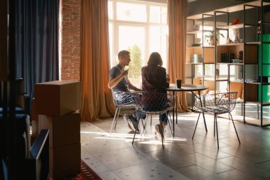 İki kişi, şık ve aydınlık bir dairede kahve içerken sohbet ediyorlar.