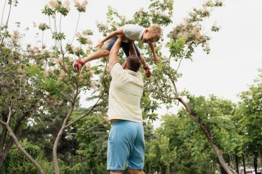 Neşeli bir baba, ağaçlarla çevrili yeşil bir park ortamında oğlunu havaya kaldırarak bir mutluluk anı yakalıyor ve bağ kuruyor..