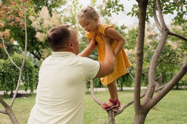 Baba, güneşli bir günde parkta ağaca tırmanırken küçük kızına yardım ediyor. Aile bağlarını ve desteğini vurguluyor..
