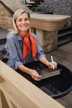 Kırmızı eşarplı neşeli bir kadın açık bir kafede otururken dijital tablete yazıyor. Gündelik ve modern bir ortam yaratıcılık ve rahatlama hissi uyandırıyor..