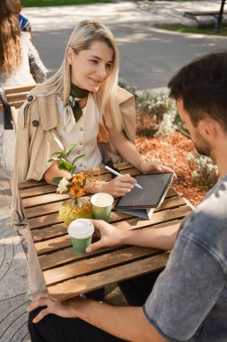 İki kişi açık bir kafede dostça bir iş görüşmesi yapıyor. Sahne bir tablet ve kahve içeriyor, rahat bir ortamda üretkenlik ve gündelik konuşma öneriyor..