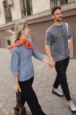 Bir şehir caddesi boyunca el ele gezen neşeli bir çift, keyifli bir günün tadını çıkarıyor. Gülüşleri ve günlük kıyafetleri mutluluğu ve güçlü bir bağı yansıtıyor..