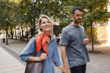 Neşeli bir çift, dışarıda güneşli bir günün tadını çıkarıyor, el ele tutuşuyorlar ve yeşil ağaçlar ve sokak lambaları arasında gülümsüyorlar. Aşk, mutluluk ve rahatlık kavramları için mükemmel..