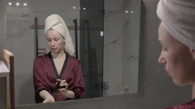 Kafasında havlu olan bir kadın, bornoz giyiyor, modern banyo aynasına parfüm sürüyor..