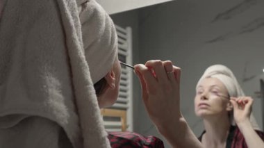 Kafasına havlu sarılmış bir kadın banyodaki aynanın önünde fırçayla kaşlarını tarıyor..