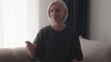 Genç Yetişkin Kadın Televizyon İzlemek İçin Uzaktan Kumanda Kullanıyor. Televizyon izleyen, kanal değiştiren ve koltukta dinlenen bir kadın..