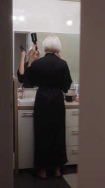 Kısa beyaz saçlı bir kadın modern bir banyoda aynanın önünde saçını tarıyor. O uzun siyah bir ipek bornoz giyiyor..