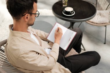 Bir adam açık bir kafede rahatça oturup deftere bir şeyler yazıyor. Dekor rahat ve kahve fincanlı siyah bir masa içeriyor. Yaratıcılık ve ilham kavramları için ideal..