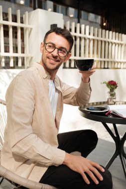 Bardaklı bir adam, güneşli bir günde mutluluk ve rahatlama saçan açık hava bir kafede bir fincan kahvenin tadını çıkarır. Modern ve davetkar bir ortam..