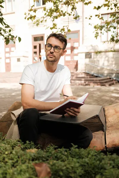 Gözlüklü düşünceli bir adam dışarıda oturur ve bir deftere yazar. Güneşli ortamda ağaçlar ve arka planda bir bina var. Huzurlu ve düşünceli bir atmosfer yaratıyorlar..