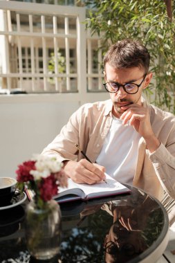 Kafe masasında oturan gözlüklü bir adam, düşünceli bir şekilde deftere yazıyor. Yakınına bir fincan kahve ve çiçek yerleştirilir, rahat bir açık hava ortamı yaratır..