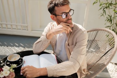 Bir adam açık bir kafede oturur ve deftere yazarken düşünür. Bir fincan kahve ve sakin bir atmosfer odaklanmasını sağlar..