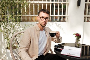 Bir adam açık bir kafe masasında oturuyor, not alırken kahvenin tadını çıkarıyor. Ortam, bitkiler ve doğal ışıkla gevşetilmiş bir atmosferi içeriyor..