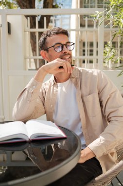 Düşünceli bir adam açık bir masada oturur, güneşli bir günde açık bir kitapla bir anlık düşüncenin tadını çıkarır. Rahatlama ve içgözlem yayar..