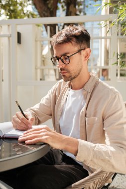 Gözlüklü genç bir adam açık bir kafe masasında otururken bir deftere yazar. Parlak atmosfer odaklanmayı ve yaratıcılığı gösteriyor..