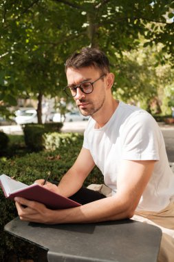 Gözlüklü bir adam dışarıda güneşli bir günün tadını çıkarırken bir not defterine yazıyor. Etrafı yemyeşil ağaçlarla çevrili, düşünceli ve rahat görünüyor..