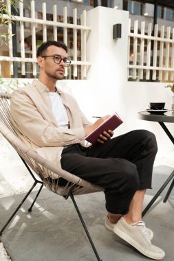 Düşünceli genç bir adam dışarıda oturur, günlük giysiler giyer, bir deftere yazarken. Yanında bir kahve fincanı ile rahat ve düşünceli görünüyor. Huzurlu ve üretken bir an olduğunu gösteriyor..