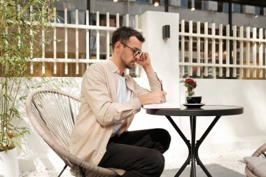 Gözlüklü şık bir genç adam açık bir kafeterya masasında deftere yazıyor. Ortam rahat ve odaklanmış, doğal ışık hoş bir ortam yaratıyor..