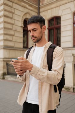 Şehir ortamında genç bir adam, akıllı telefonuna dikkatle bakıyor. Sıradan giyinmiş ve bir sırt çantası takıyor. Seyahat ya da keşif teması öneriyor..