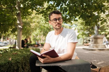 Parktaki bankta oturan genç bir adam güneşli bir günde kırmızı bir günlük yazıyor. Etrafı yeşilliklerle çevrili, bu sakin açık havada düşünceli ve rahat görünüyor..