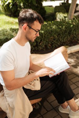 Genç bir adam parkta bir bankta oturuyor, bir kitap okumaya dalmış. Açık hava manzarası güneşli ve huzurlu, yeşilliğin ortasında bir anlık rahatlama ve odaklanma sunuyor..