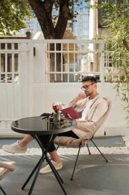 Gündelik kıyafetli genç bir adam açık bir kafe masasında oturuyor elinde bir defter, derin düşünceler içinde. Sahne huzurlu ve kentsel, rahatlık ve tefekkürün birleşimi..