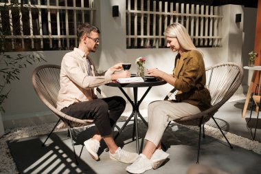 Bir adam ve kadın bir kafede karşılıklı oturup kahvenin tadını çıkarıyorlar. Dış mekan güneş ışığıyla yıkanır, rahat ve dostane bir atmosfer yaratır..