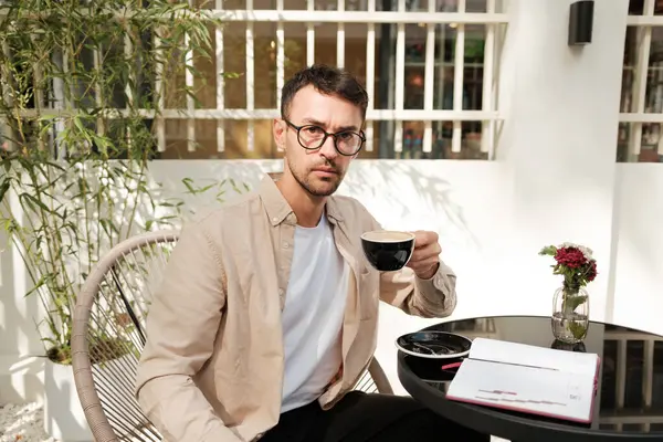 Bir adam açık bir kafe masasında oturuyor, not alırken kahvenin tadını çıkarıyor. Ortam, bitkiler ve doğal ışıkla gevşetilmiş bir atmosferi içeriyor..