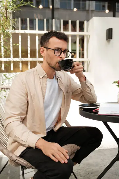 Genç bir adam açık bir kafede oturuyor, kahvesini yudumluyor ve dinleniyor. Gelişigüzel giyinmiş, sakin ve sade bir yaşam tarzını somutlaştırıyor, şık bir şehir zeminine dayanıyor..