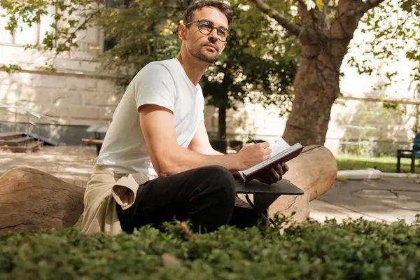 Gözlüklü düşünceli bir adam parkta bir kütüğe oturur, dikkatle defterine yazar, yeşillik ve güneş ışığıyla çevrilidir. Sahne yaratıcılık, yansıma ve rahatlama içerir..