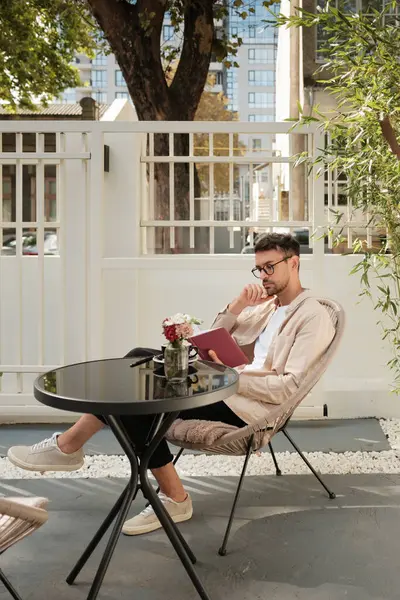 Gündelik kıyafetli genç bir adam açık bir kafe masasında oturuyor elinde bir defter, derin düşünceler içinde. Sahne huzurlu ve kentsel, rahatlık ve tefekkürün birleşimi..