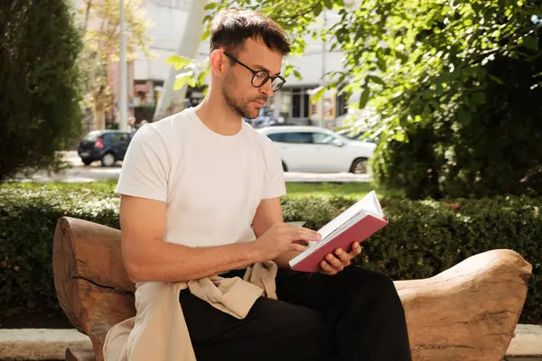 Güneşli bir parktaki ahşap bir bankta oturan bir adam kitap okumaya odaklanmış. Etrafı yeşilliklerle çevriliyken, huzurlu bir rahatlama ve yalnızlık anının tadını çıkarıyor..