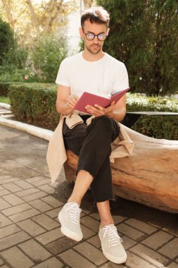 Gözlüklü genç bir adam güneşli bir bahçedeki ahşap bir bankta oturuyor, bir kitap okumaya dalmış. Rahatlamış ve odaklanmış görünüyor. Açık hava ortamının tadını çıkarıyor..