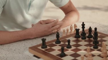 Bir satranç oyununda bir sonraki hamlesini düşünen bir adamın ellerine yaklaş. Kamera ellerine ve satranç tahtasına odaklanıyor. Oyunda gereken stratejik derinliği ve konsantrasyonu vurguluyor..