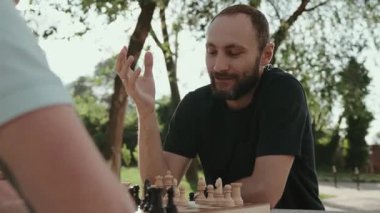 İki erkek arkadaş güneşli bir günde parkta satranç oynuyorlar. Gülümsüyorlar ve oynarlarken konuşuyorlar..