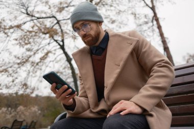 Bir bankta oturmuş açık havada akıllı telefonu kontrol eden bir adam.