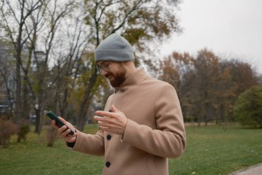 Sonbaharda parkta bir adam, palto ve bere giyiyor, telefonu tutuyor.