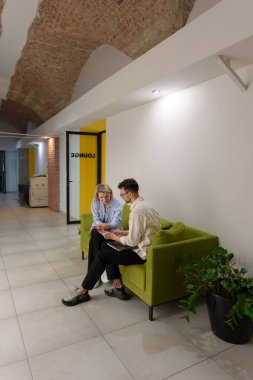 İki kişi bir koltukta oturmuş, bir ofis salonunda sohbet ediyorlar..