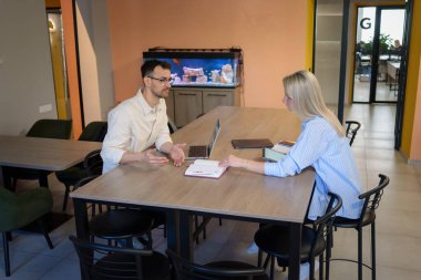 İki iş arkadaşı bir masada oturmuş, parlak bir ofiste tartışıyorlar..