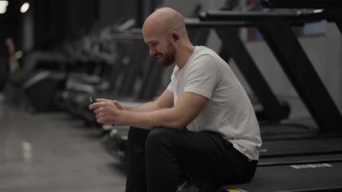 Akıllı telefon kullanan ve spor salonunda gülümseyen bir adam. İletiler, sosyal medya kontrol ediliyor.