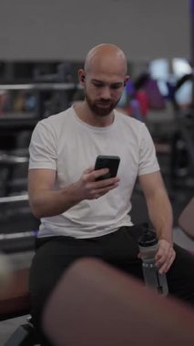 Kel adam spor salonunda oturur, telefonu kontrol eder ve suyu yudumlar..