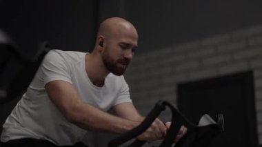 Spor salonunda spor bisikletini kullanan kel sakallı adam..