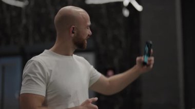 Sakallı kel adam spor salonunda selfie çekiyor.