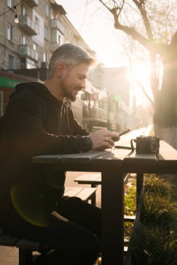 Güneşli bir günde bir adam kameralı telefonuyla dışarıda oturur..