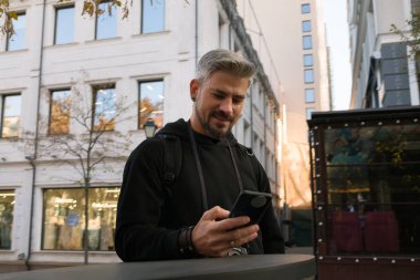 Bir adam şehir sokağında dikilirken akıllı telefonuna bakar..