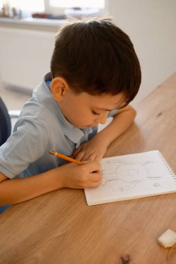 Genç bir çocuk parlak ahşap bir masada bir defter çizerken konsantre olur. Sahne yaratıcılığın özünü yakalar ve sakin bir iç mekanda odaklanır..