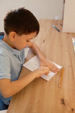 Genç bir çocuk ahşap bir masadaki defterde bir kalemle çizim yapmaya konsantre oluyor. Resim, çocukluk öğrenimi ve sanatsal ifadeyi vurgulayarak yaratıcılık ve odaklanma anını yakalar..