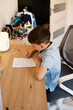 Genç bir çocuk masasında bir resim çizmeye odaklanmış. Masa renkli yapı taşları ile organize edilir ve yaratıcı bir atmosfer sağlar, hayal gücü ve öğrenme faaliyetleri için idealdir..